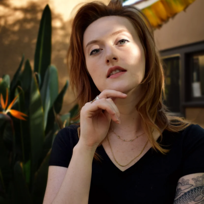 Photo of Cassidy Pearsall, a white woman with red hair wearing a black short-sleeved v-neck shirt and red lipstick, holding her hand up to her chin.