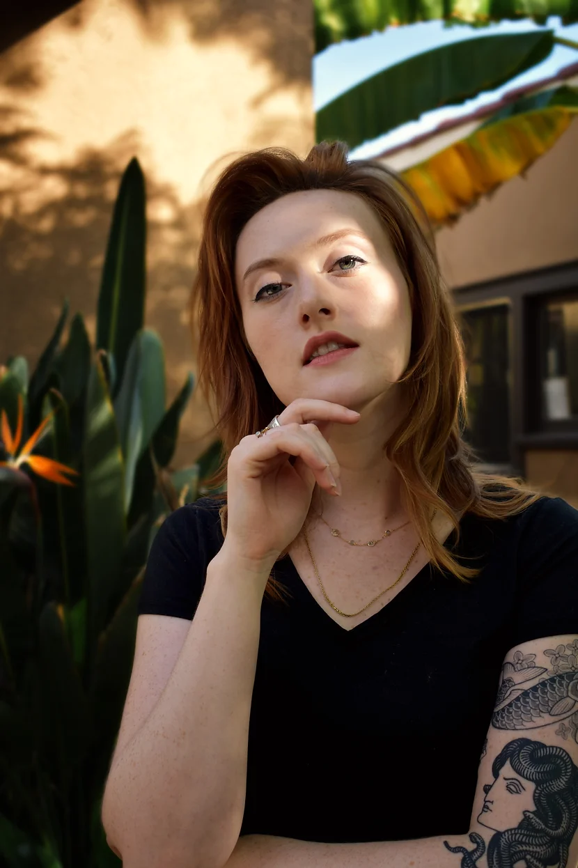 Photo of Cassidy Pearsall, a white woman with red hair wearing a black short-sleeved v-neck shirt and red lipstick, holding her hand up to her chin.