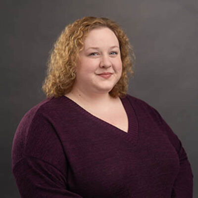 Photo of Cat Wilson, a white woman with blonde curly hair, wearing a deep purple v-neck sweater