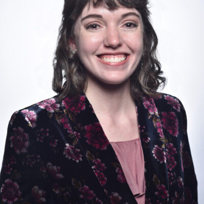 Photo of Celia Kasberg, a woman with brown hair and bangs, wearing a flower patterned velvet jacket.