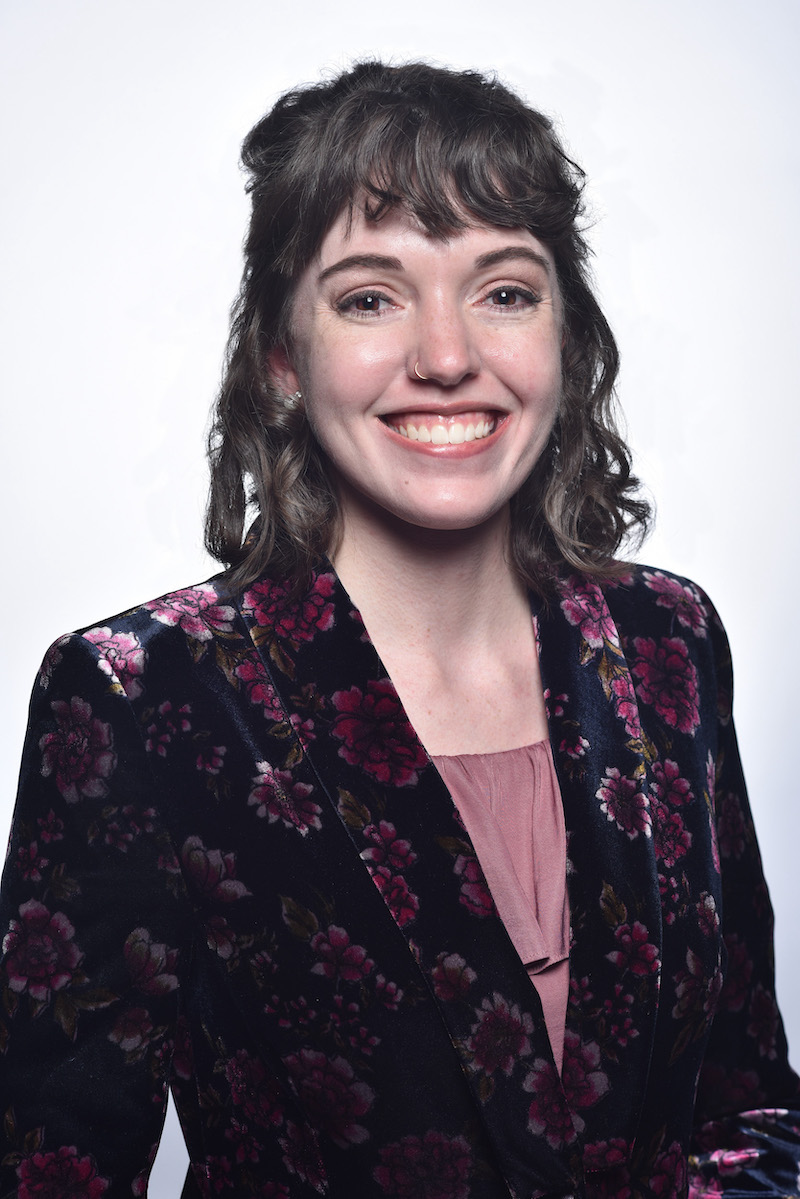 Photo of Celia Kasberg, a woman with brown hair and bangs, wearing a flower patterned velvet jacket.