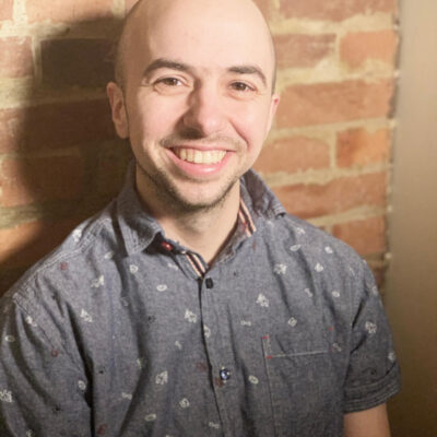 Photo of Chase Trumbull, a white man with a bald head, wearing a grey short-sleeved button up shirt, leaning against a brick wall.