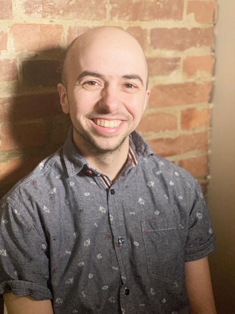 Photo of Chase Trumbull, a white man with a bald head, wearing a grey short-sleeved button up shirt, leaning against a brick wall.