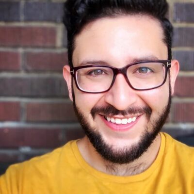 Photo of Damian Dominguez, a man with dark brown hair and short beard, wearing glasses and a bright yellow shirt, a brick wall in the background.