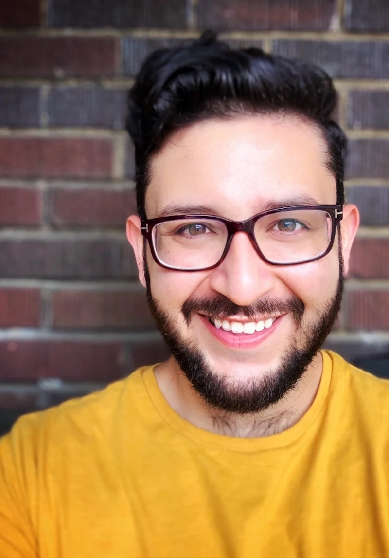 Photo of Damian Dominguez, a man with dark brown hair and short beard, wearing glasses and a bright yellow shirt, a brick wall in the background.