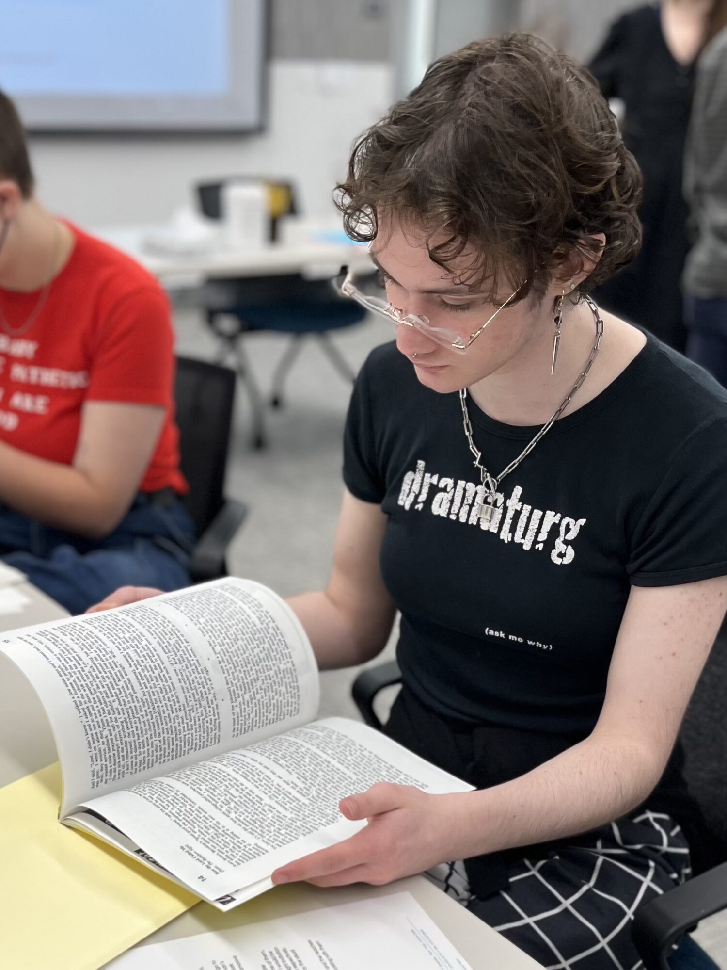 A student wearing a black t-shirt that says "dramaturg" in white letters, sits at table in a classroom, reading a book.