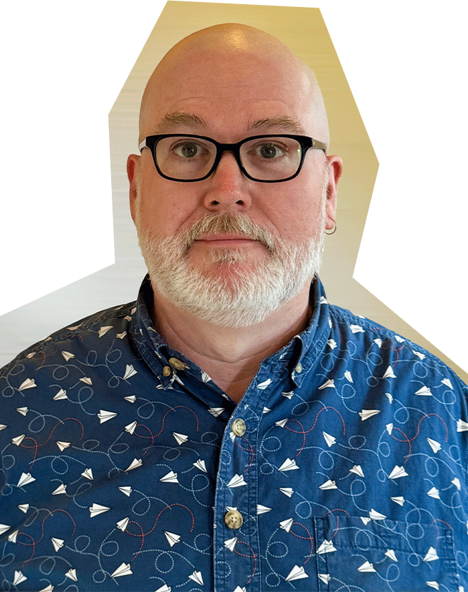 Photo of David Holcomb, a white man with a bald head and white facial hair, wearing glasses and a blue collared shirt with a paper airplanes patterns.