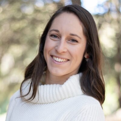 Photo of Emma Present, a woman with medium length dark brown hair, outdoors wearing a white sweater.