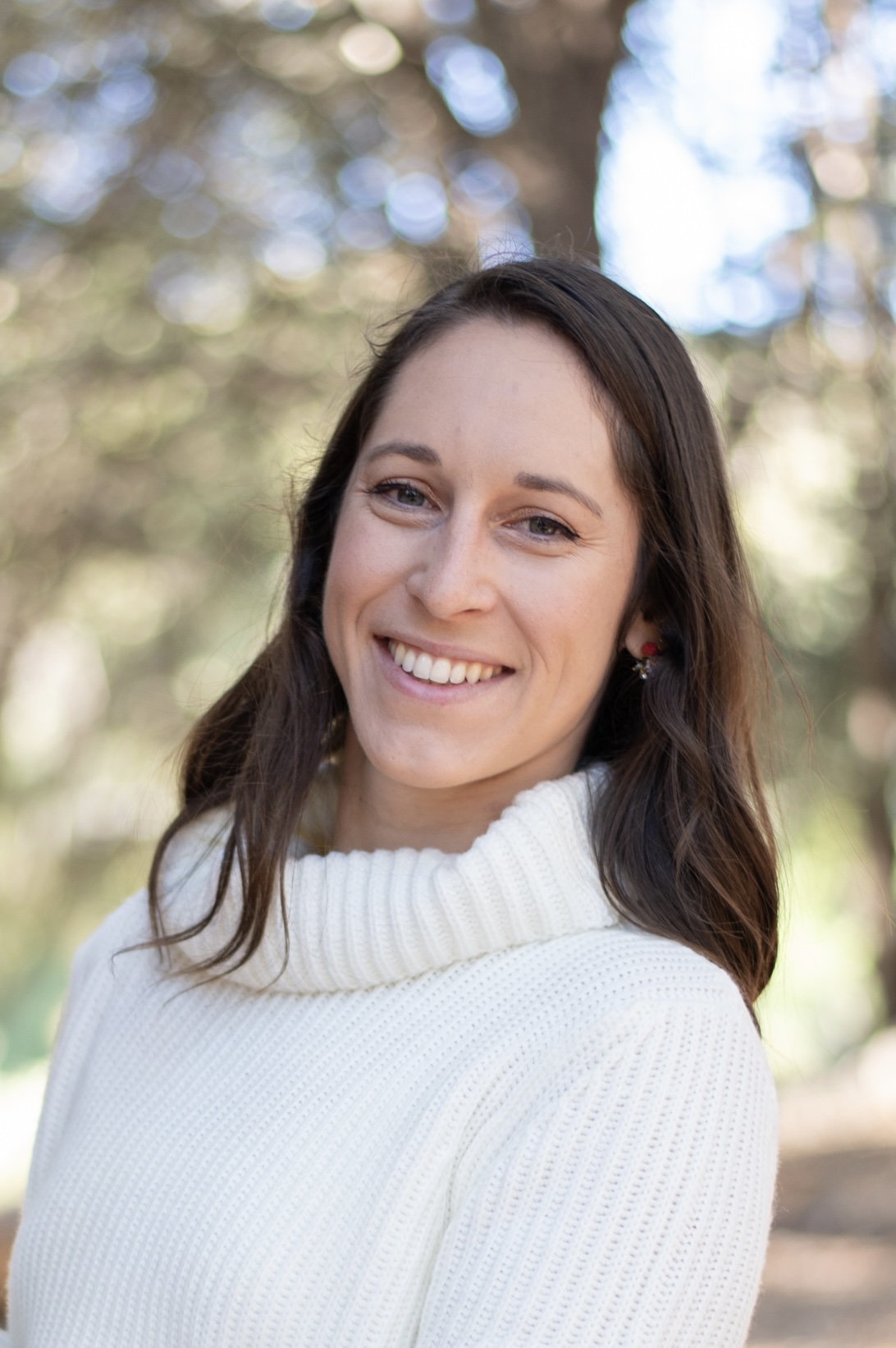 Photo of Emma Present, a woman with medium length dark brown hair, outdoors wearing a white sweater.