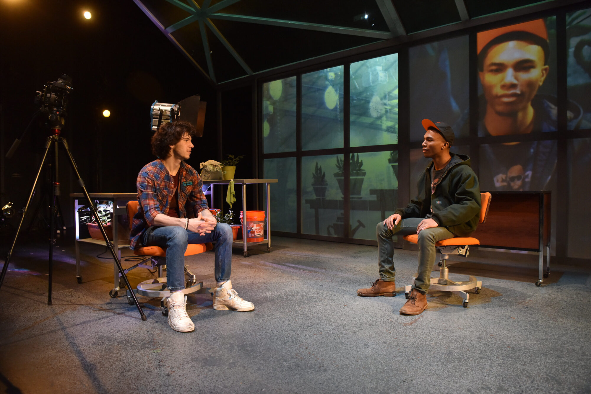 Two student actors sit in chairs on a set of a play, with projections on the wall behind them.