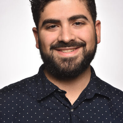 Photo of James Gallo, a man with dark curly hair and dark facial hair, wearing a navy button up shirt with small white dots.