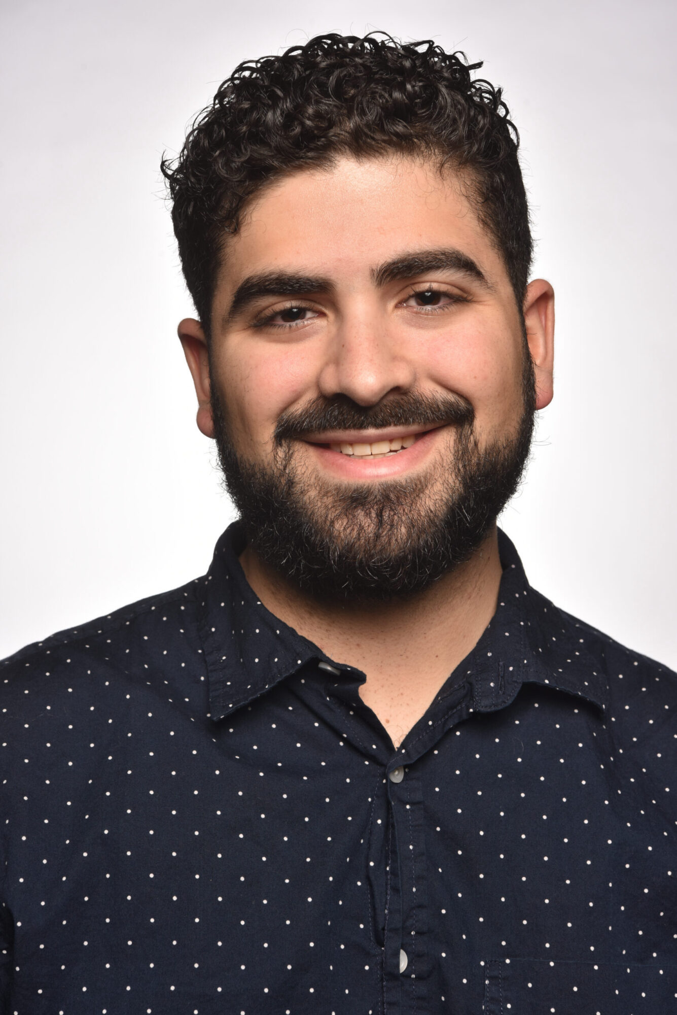 Photo of James Gallo, a man with dark curly hair and dark facial hair, wearing a navy button up shirt with small white dots.