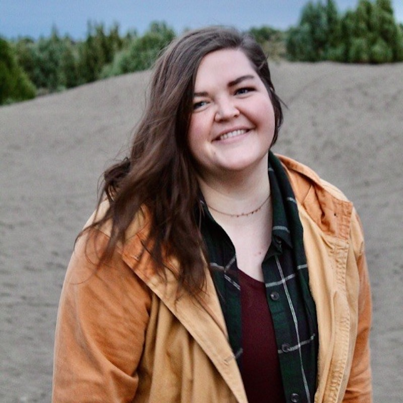 Photo of Kyrie Bayles, a white woman with long brown hair standing outside and wearing a flannel shirt and yellow jacket.