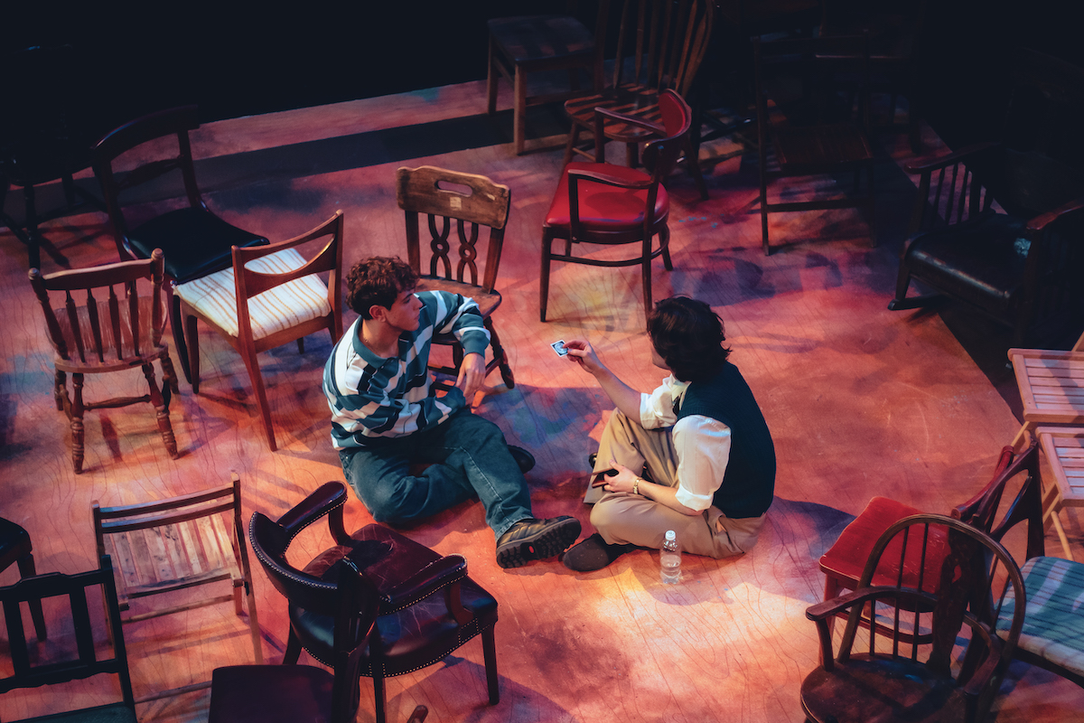 Two actors sit on the floor of a stage, surrounded by many wooden chairs.