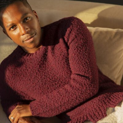 Photo of Leslie Odom, Jr. - a black man wearing a long sleeved maroon shirt and lounging on a cream colored couch.