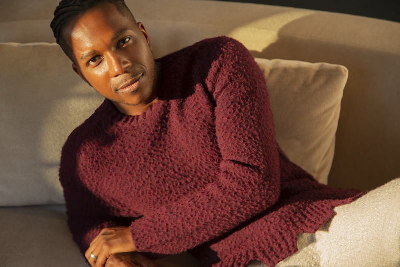 Photo of Leslie Odom, Jr. - a black man wearing a long sleeved maroon shirt and lounging on a cream colored couch.