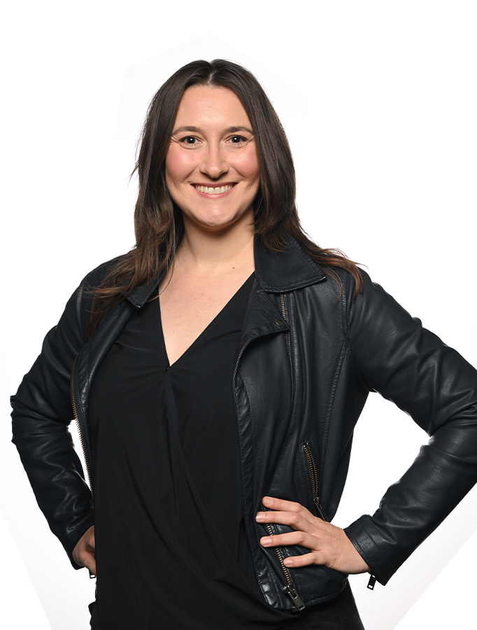 Photo of Melissa Lubina, a white woman with long dark hair wearing all black, standing with her hands on her hips and smiling.