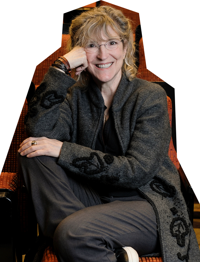 Catherine Moore, a white woman with dirty blonde hair and bangs, wearing glasses and a gray suit, sits in theater seating.