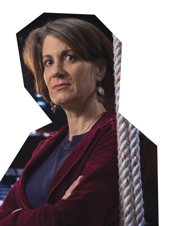 Photo of Anne Mundell, a woman with brown hair wearing a dark maroon jacket, crossing her arms, standing in the wings of a theater.