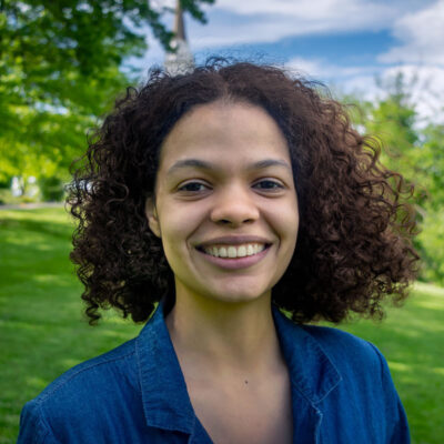 Photo of Paloma Sierra, a woman with dark curly hair, wearing a blue top, standing on a green lawn with trees behind her.