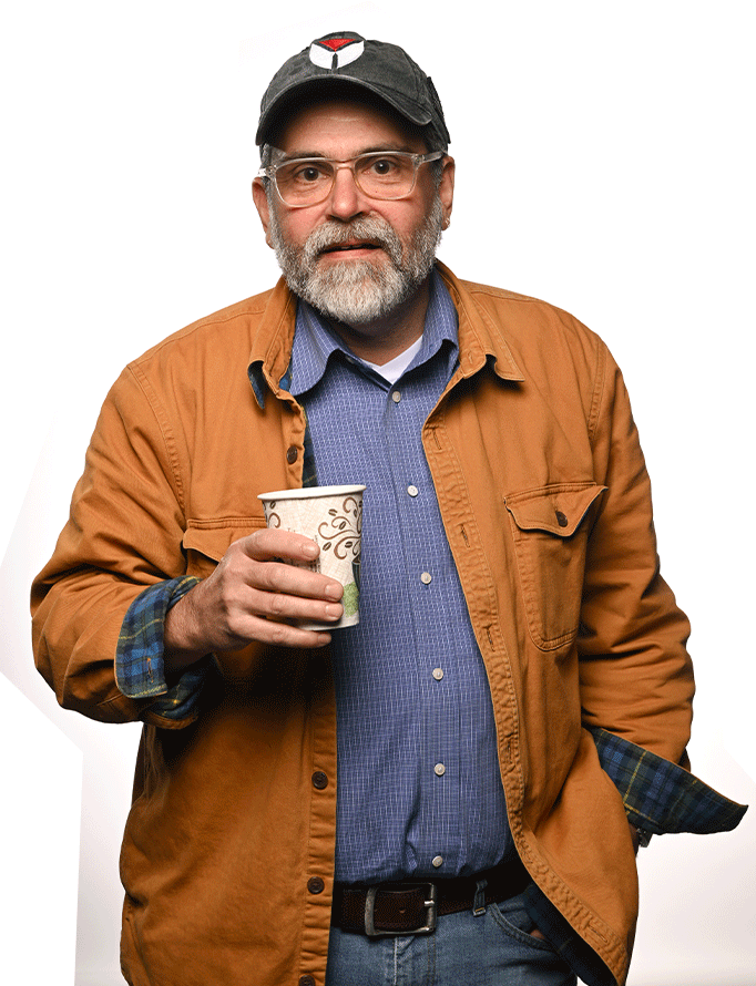 Photo of David Randolph, a white man with a beard and glasses, wearing a ball cap, blue button down shirt, and tan jacket, holding a cup of coffee.