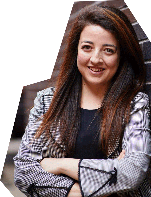 Photo of Sarah Shechtman, a white woman with long brown hair, wearing a great jacket and crossing her arms, smiling.