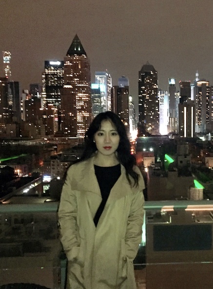 Photo of SooA Kim, a woman wearing a tan trench coat standing with a cityscape in the background.