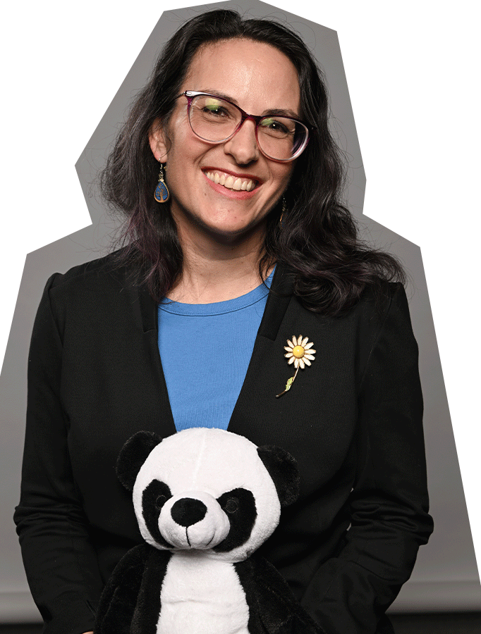 Photo of Kristin Ward, a white woman with brown hair and glasses, wearing a blue shirt and black blazer, and holding a stuffed panda.