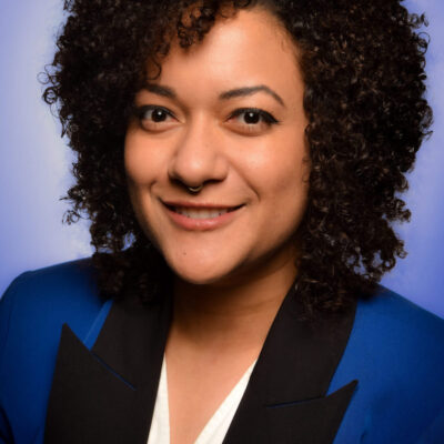 Photo of Xotchil Musser, a woman with short black curly hair wearing a blue blazer with a black lapel.