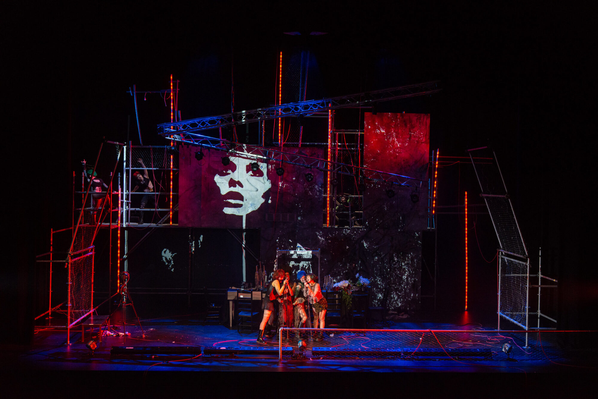 Photo of a stage with an industrial looking set lit in red with a projection of a face on one of the walls. Four actors sing into a mic on stage.