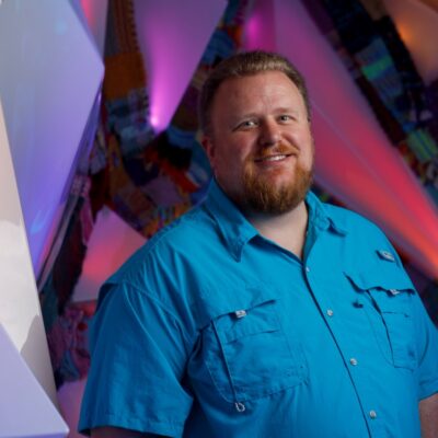 Photo of Derek Pendergrass, a man wearing a bright blue shirt standing in front of a colorfully lit background.