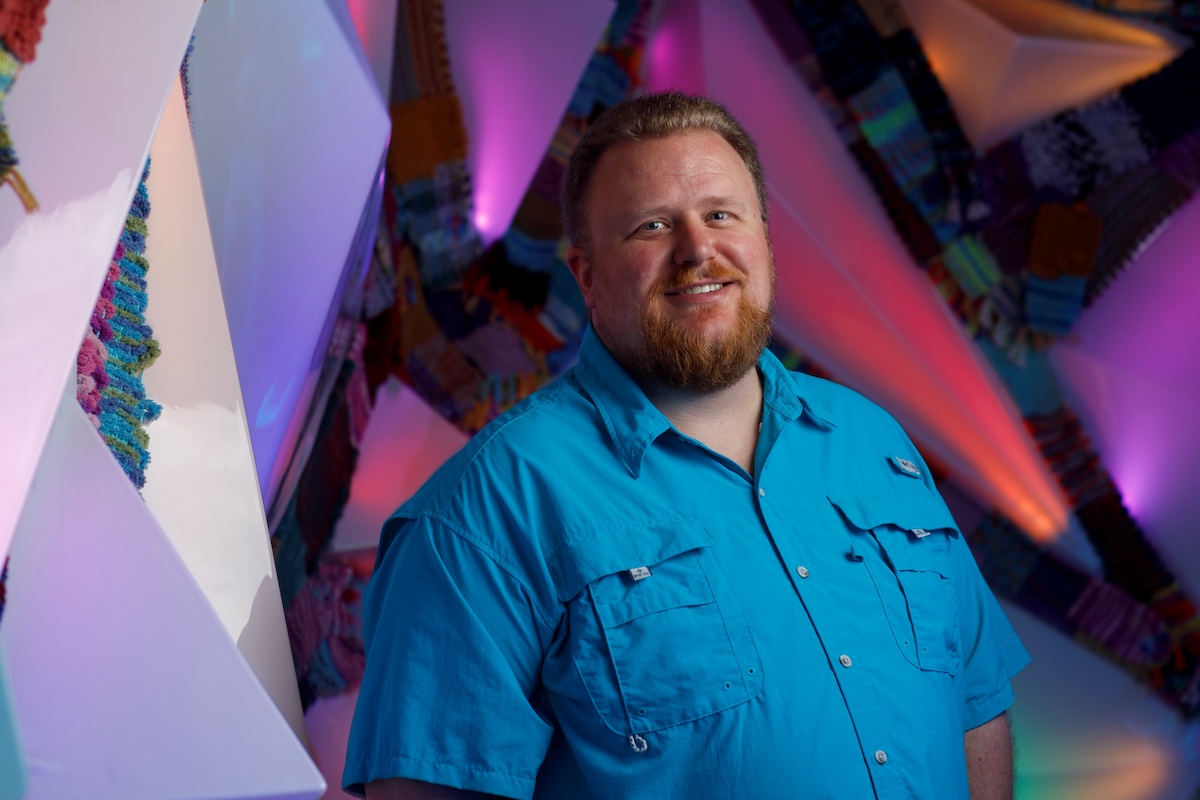 Photo of Derek Pendergrass, a man wearing a bright blue shirt standing in front of a colorfully lit background.