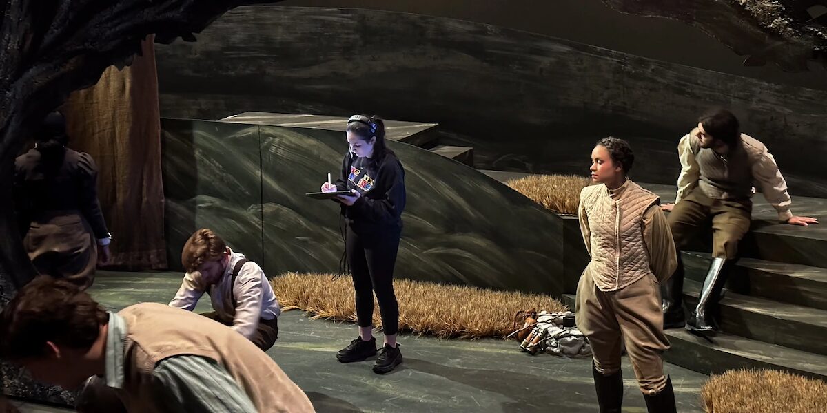 Students on stage during a rehearsal, one student in the center wears a headset and takes notes on a clipboard.