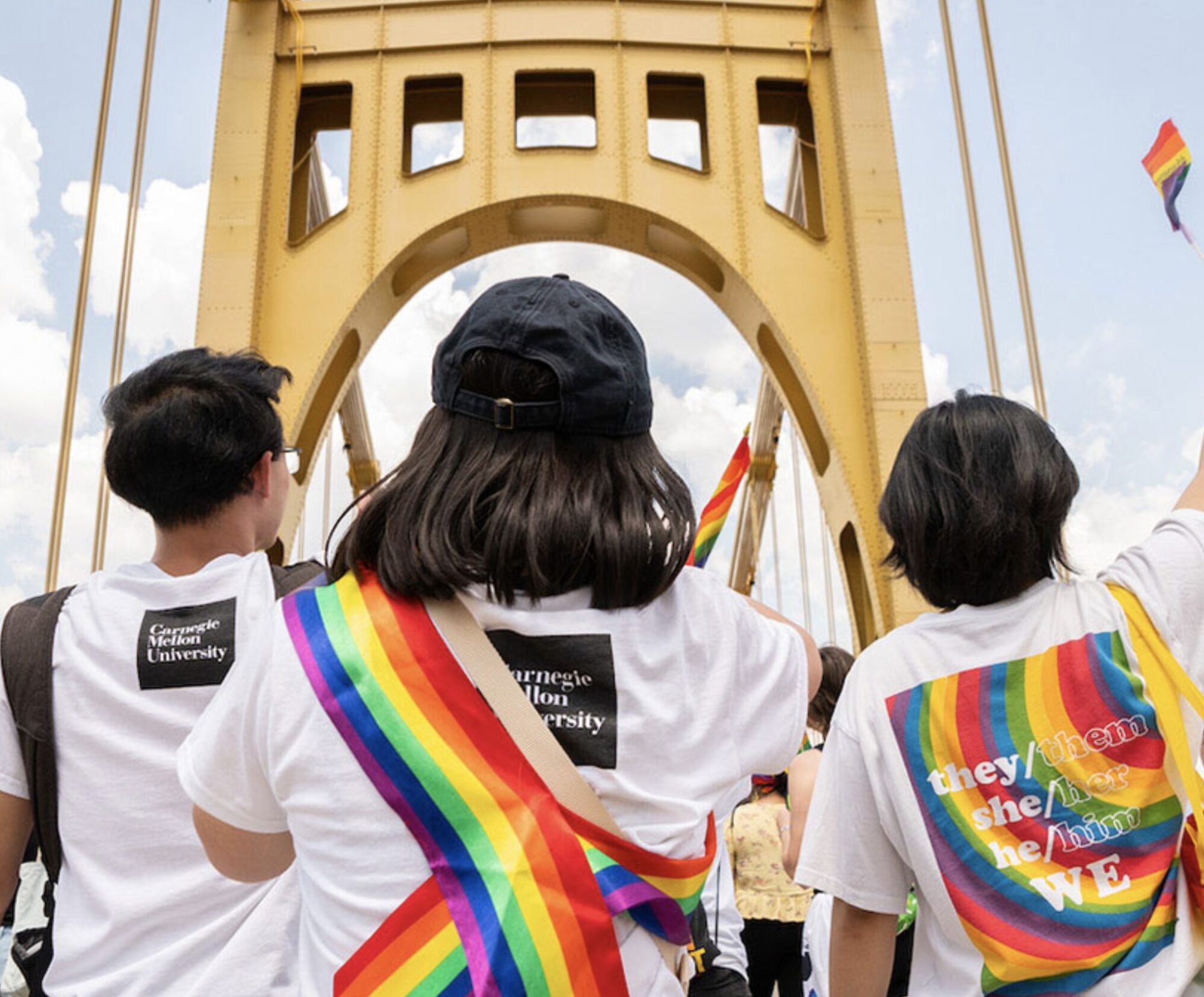 Photo of the backs of three students wearing rainbow sashes and looking up at one of Pittsburgh's yellow bridges.