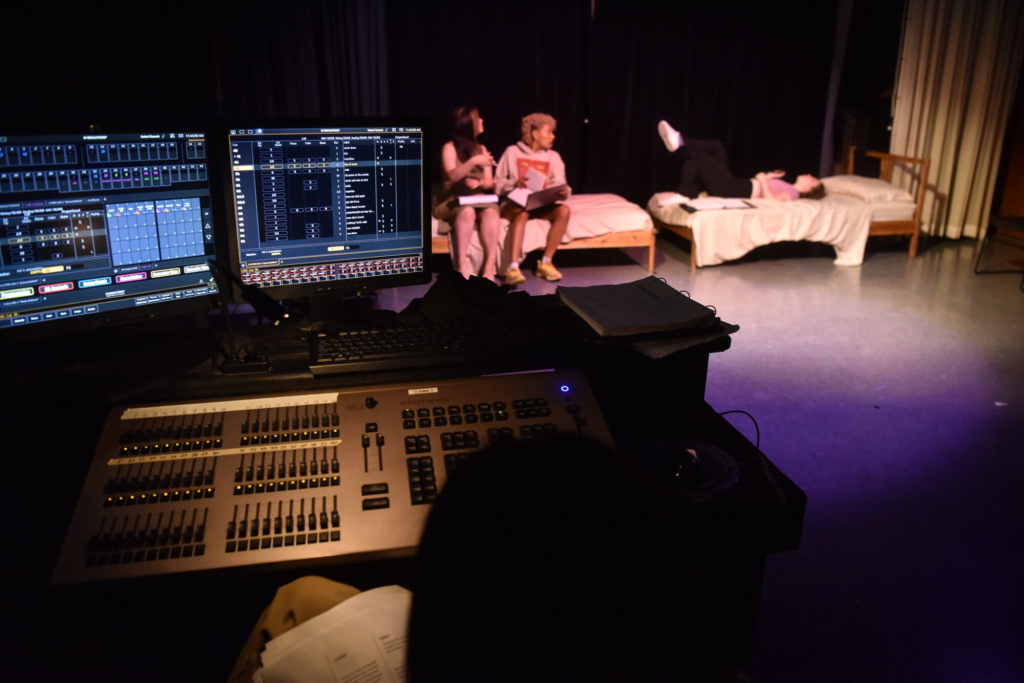 A lighting board in a dark blackbox theater; actors in the background on prop furniture.