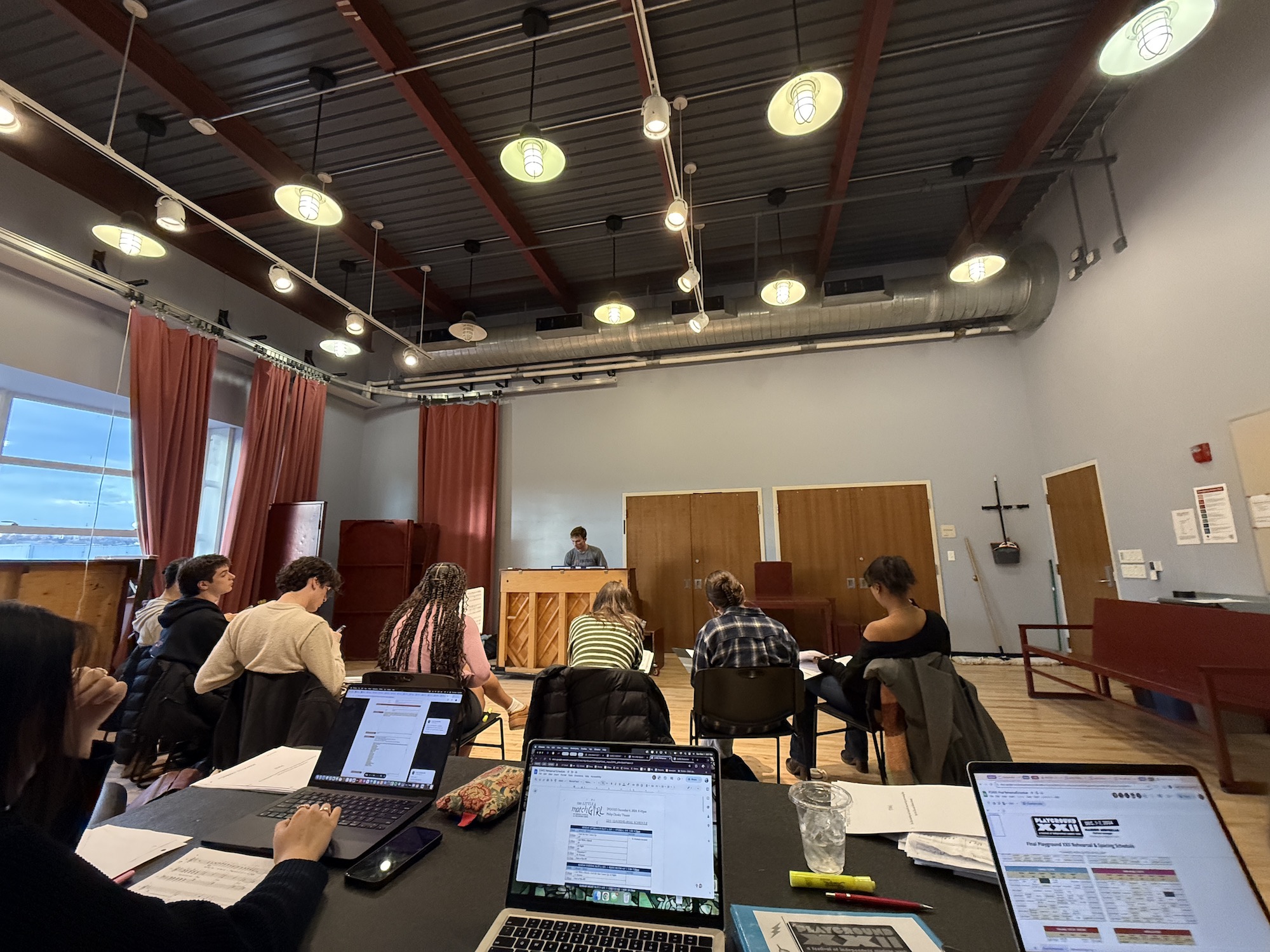 Students in a rehearsal room with laptops, some sit in chairs, one stands at a piano.