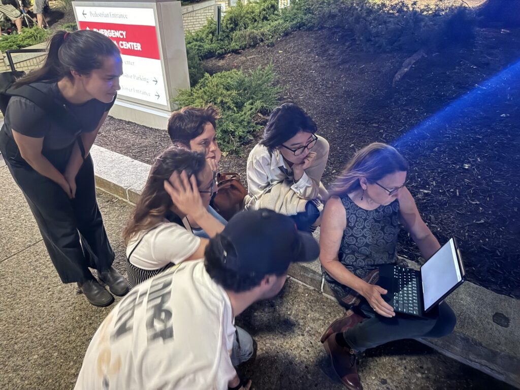 Students gather around production designer Nina Rucsio's laptop outside a Pittsburgh emergency room on a location shoot for "The Pitt."