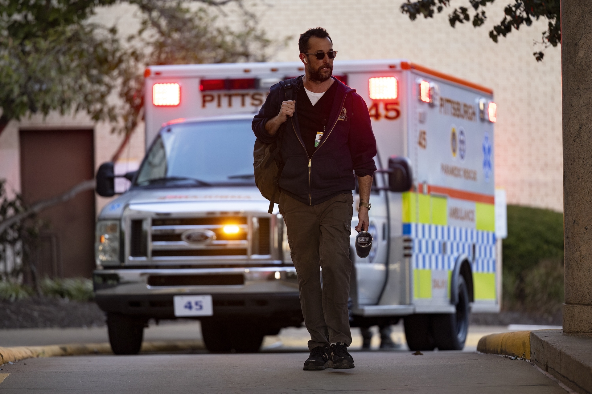 Actor Noah Wyle walks down a street, with a Pittsburgh ambulance behind him, in a scene from "The Pitt."