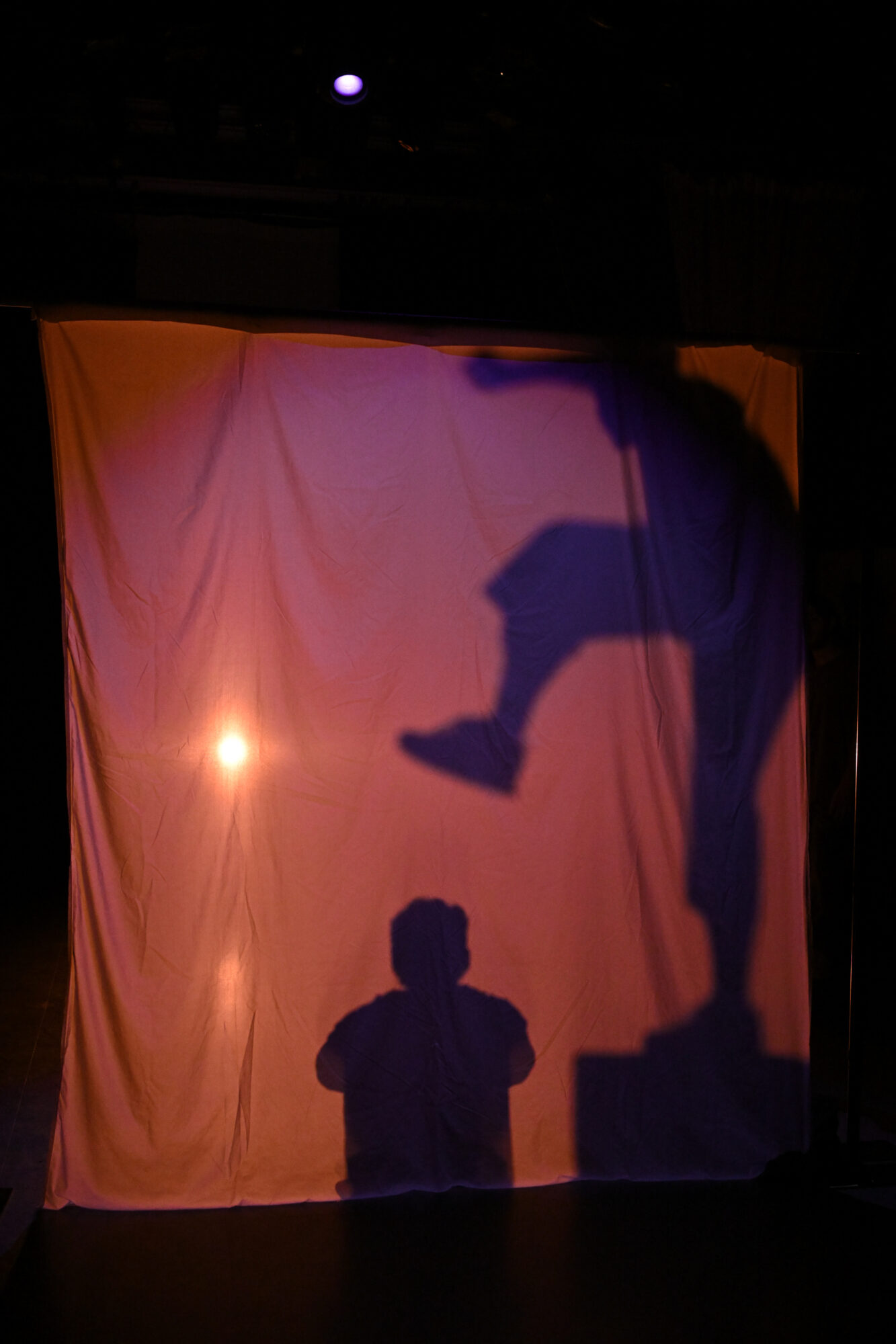 A sheet lit from behind with silhouettes of actors, one sitting appearing about to be stomped by the foot of another.
